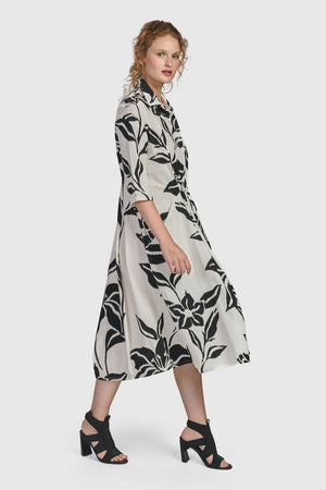 Alembika Botanica Business Dress. Black lillies print on a beige background. Pointed collar 3/4 sleeve dress with 1/2 button placket. Cinch tie at waist. Balloon skirt. Relaxed fit._34778043154632
