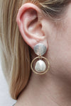 Stylish and modern dangle earrings each featuring a luminous white pearl framed by a golden brass ring hanging from a hammer textured organic shaped silver stud with a  standard hypoallergenic earring clip backing._t_33891613147336