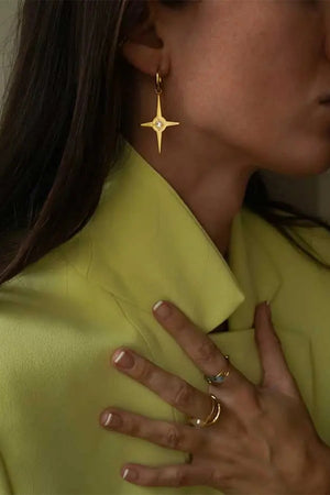 The Star Bright Earrings are eye catching and fun! These golden stainless steel earrings each feature large re-imagined star shaped pendant with a sparkling crystal rhinestone in the center hanging from standard snap huggie hoop. This elegant accessory will add interest to your outfit._33915296678088
