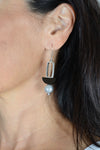 The Atmospheric Orbs Dangle Earrings are a modern and chic pair of dangle earrings each showcasing a sculptural stack of a luminous gray pearl, a black wooden half circle bead, and an open silver rectangle hanging from a standard hypoallergenic surgical steel fishhook earring wire._t_33053530751176